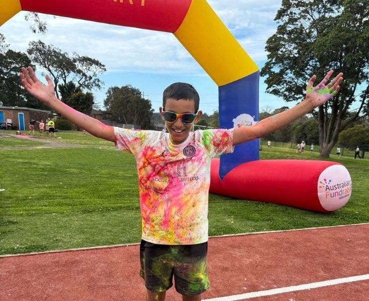 A boy wearing a white shirt covered in coloured powder. He has his hands outstretched.