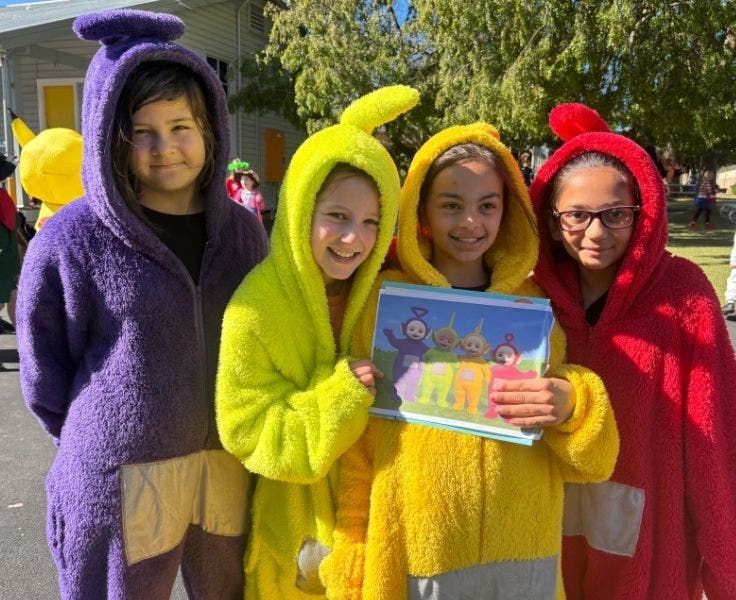 Four girls wearing teletubby costumes, one is purple, one is lime, one is yellow and one is red. They are holding a Teletubby book.