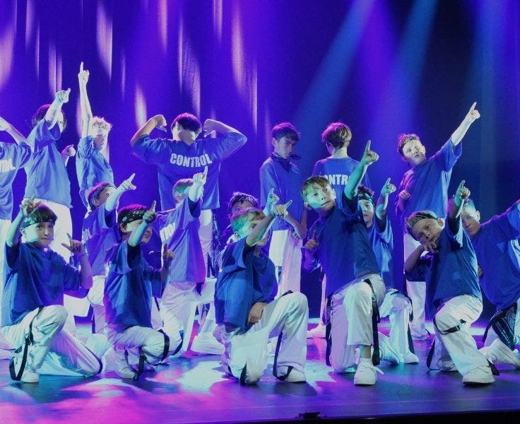 A group of boys on a stage holding a dance move. They are wearing a blue t-shirt and white pants. The back of their top has the word, control!