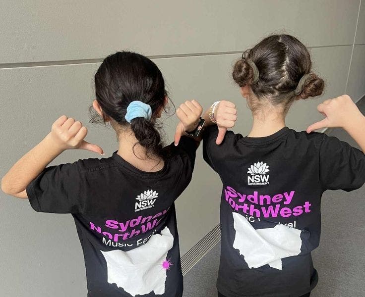 Two girls with their backs to the camera. They are wearing shirts that say Sydney NorthWest Music Festival