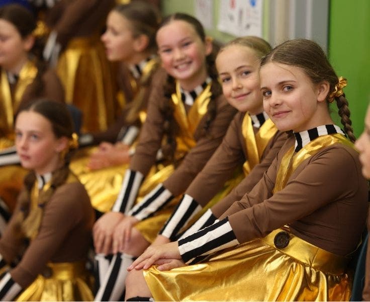 a group of girls, sitting, and wearing brown and gold dance costumes looking at the camera