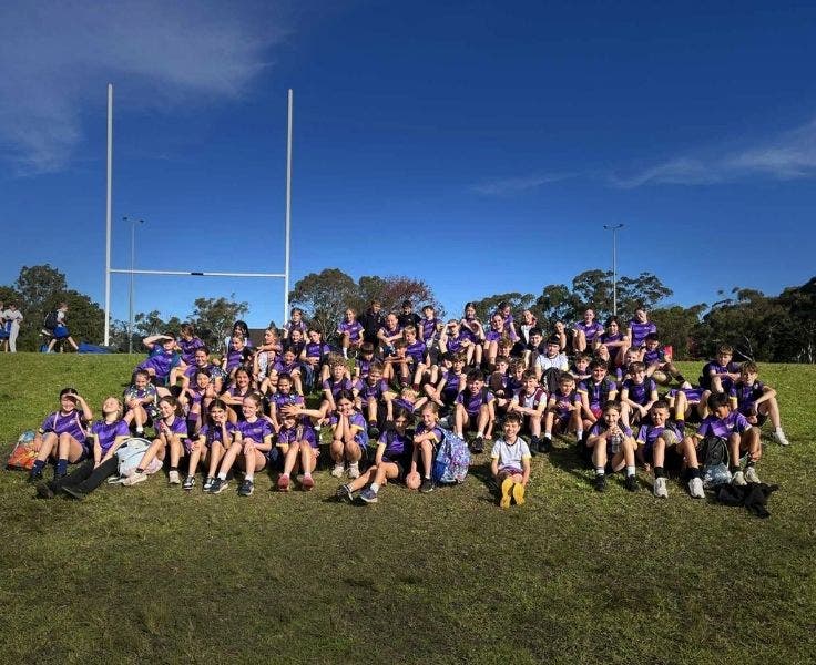 A large group of boys and girls sitting on a field with football goals behind them.